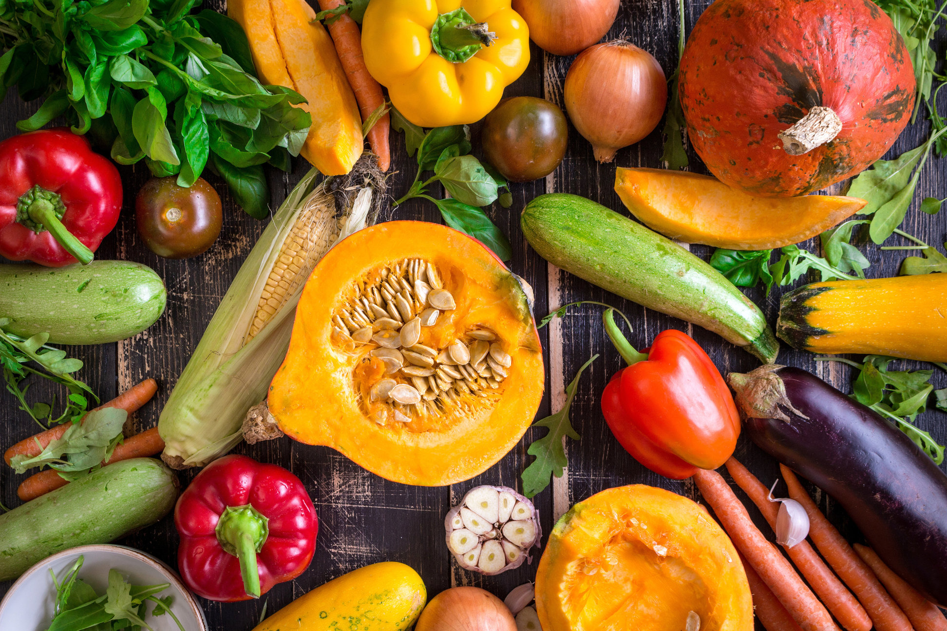 Fresh vegetables scattered on a old rustic dark textured table. Autumn background. Healthy eating. Sliced pumpkin, zucchini, squash, bell peppers, carrots, onions, cut garlic, tomatoes, eggplant, corn cob, rucola and basil. Top view